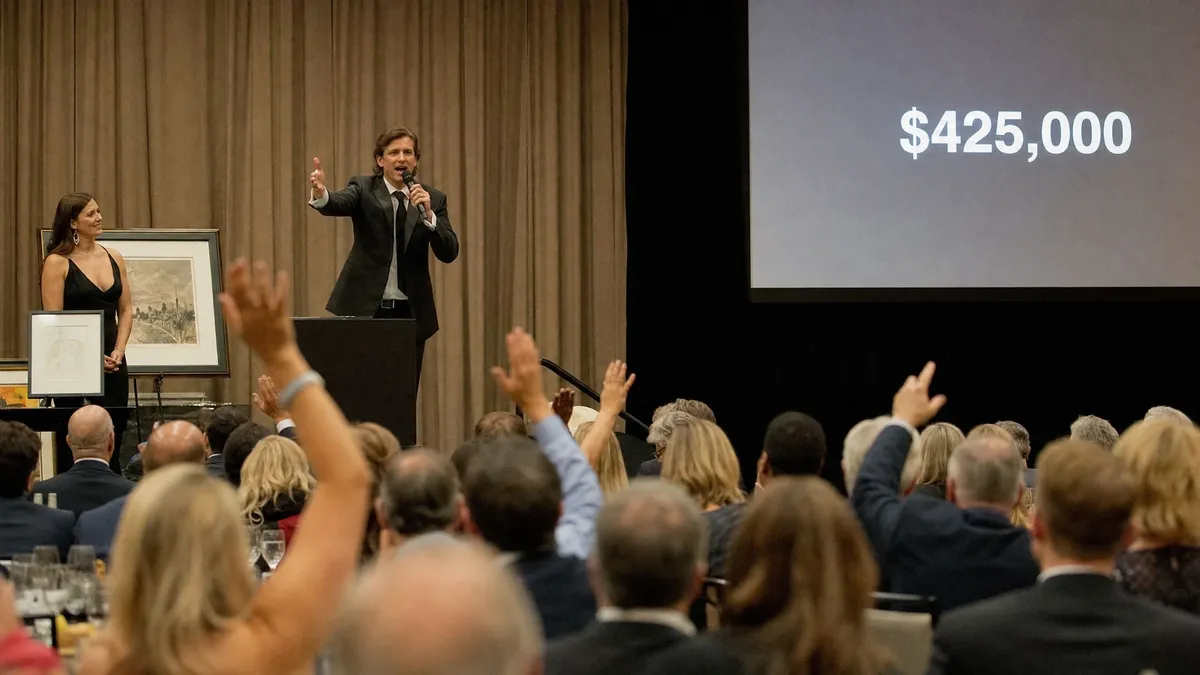 Auctioneer driving bids on a gala stage as the total climbs on screen